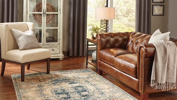 Cream, blue, and red distressed traditionally patterned rug on wood floor with brown leather couch, white fabric chair, and white shabby chic cabinet